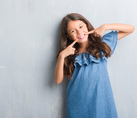 A young girl wearing a denim dress and pointing to her healthy smile