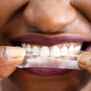 closeup of woman wearing berry-colored lipstick applying teeth whitening strip to teeth