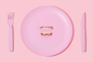 Dentures sitting on a pink plastic plate next to a pink fork and knife on a pink background